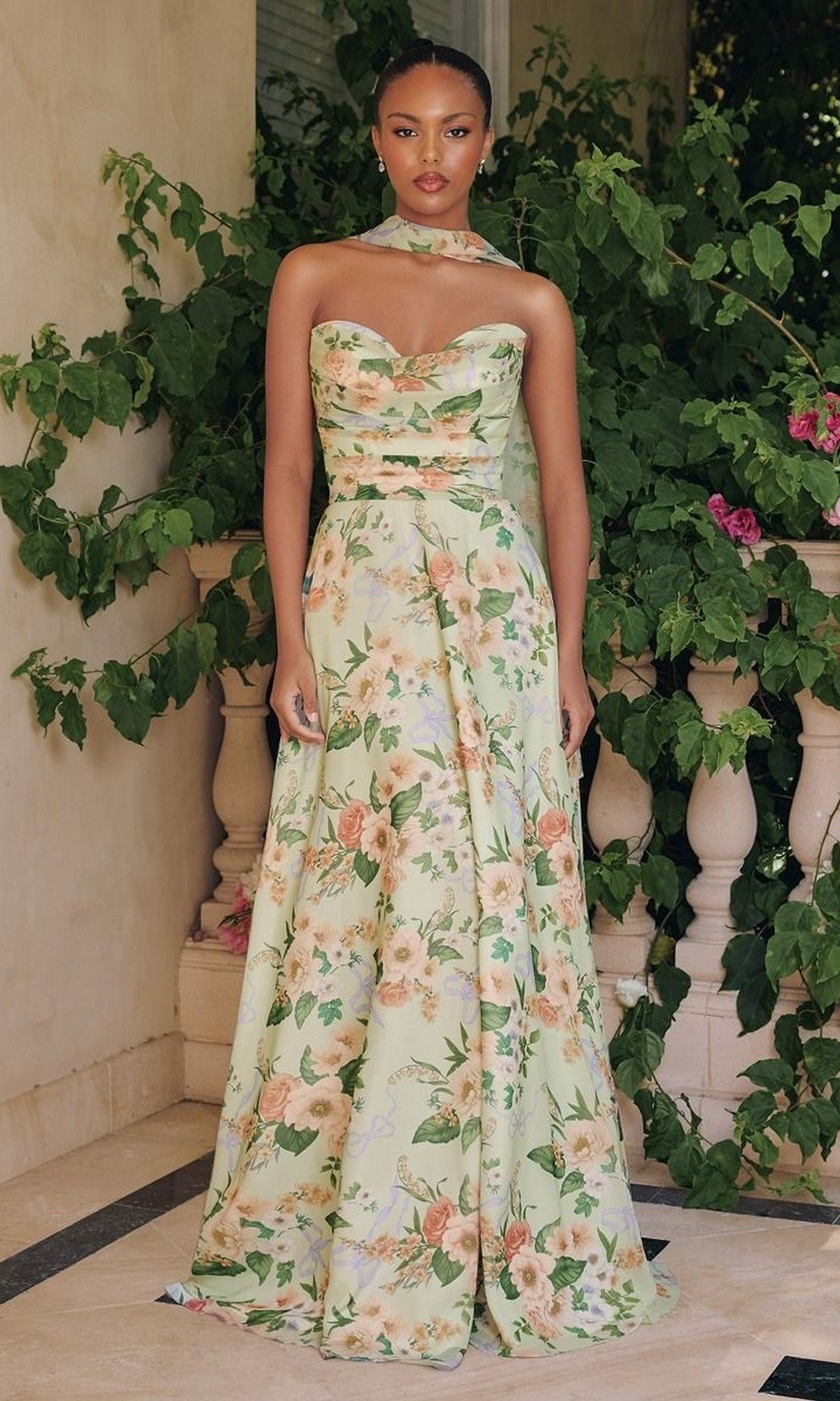 A woman poses before a leafy backdrop in the Amoris T717: a strapless, floor-length prom dress in sage print, paired with a matching neck scarf.